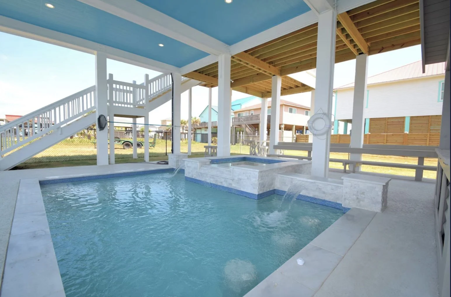 Covered backyard with swimming pool and hot tub, surrounded by a white fence, with neighboring houses visible in the background.