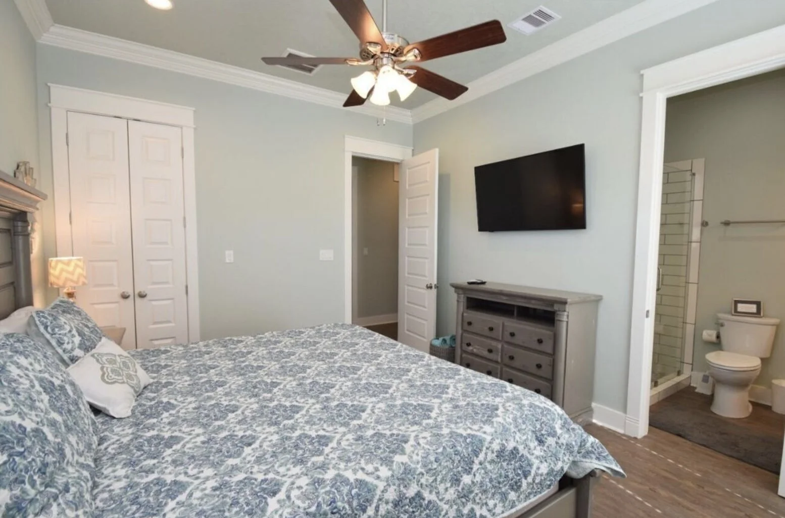 A bedroom with light blue walls, a gray wooden bed with blue patterned bedding and pillows, a nightstand with a chevron-patterned lamp, a ceiling fan with light, a flat-screen TV on the wall, a dresser, and an open door leading to a bathroom with a t