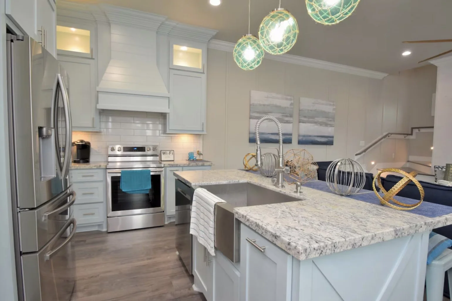 Modern kitchen with white cabinets, stainless steel appliances, granite countertops, and nautical-themed decorative objects, with a dining area and staircase in the background.