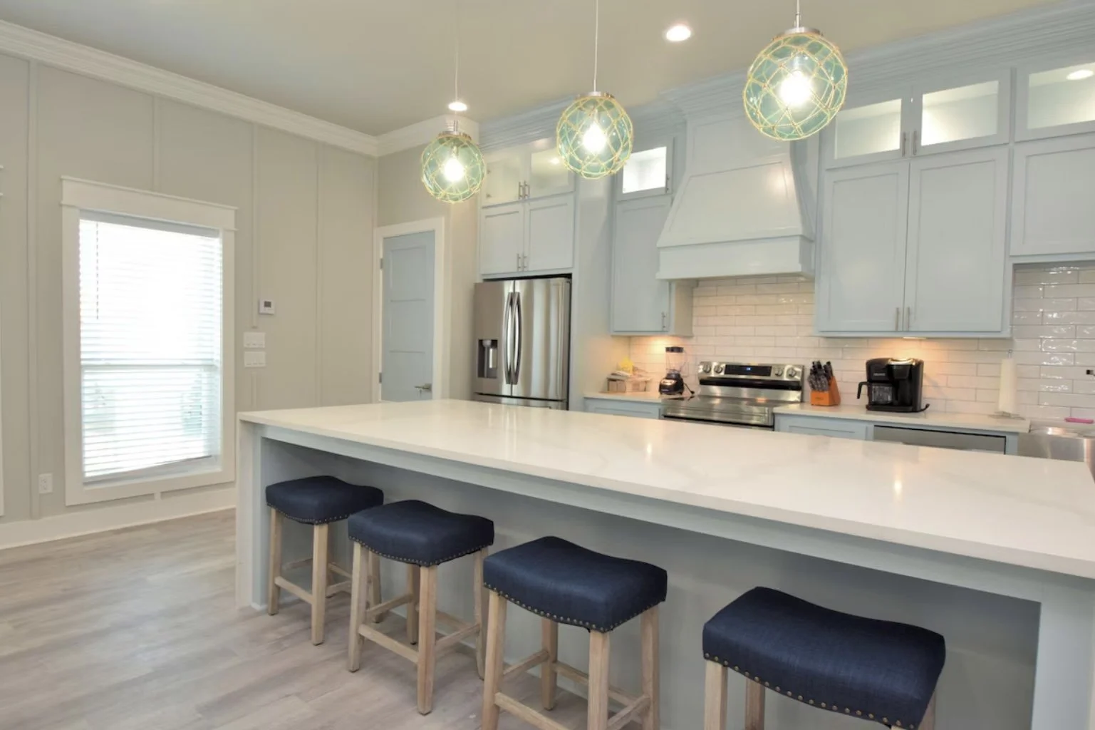 Modern kitchen with light blue cabinets, white countertops, stainless steel refrigerator, white subway tile backsplash, and four pendant lights with colorful globes over a breakfast bar with four navy stools.