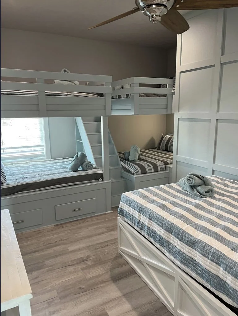 A bedroom with a bunk bed and two twin beds, all with striped bedding, and rolled towels placed on each bed. There is a window with blinds and a ceiling fan with wooden blades.