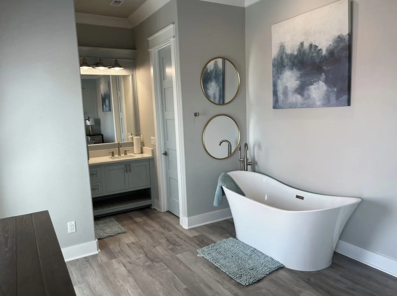 Modern bathroom with a white freestanding bathtub, two round mirrors, wall art, and a vanity with a sink, faucet, and a mirror.