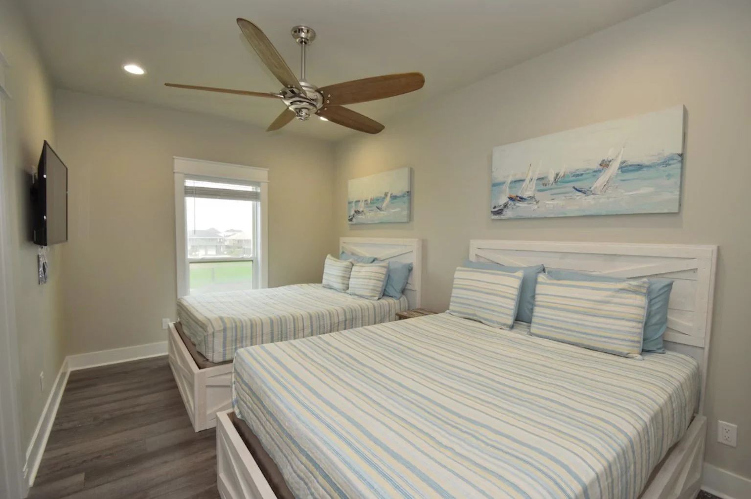 Two beds with striped bedding and pillows in a bright beach-themed bedroom. Wall art depicts sailboats on the water. There is a window letting in natural light and a ceiling fan overhead.