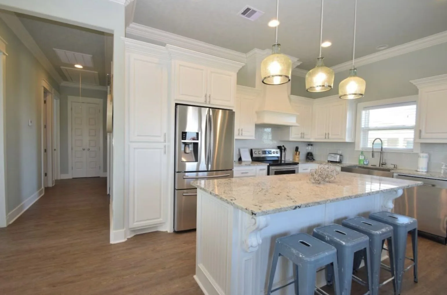 Modern kitchen with white cabinets, stainless steel refrigerator and appliances, granite countertop island, wood flooring, pendant lights, and a window above the sink.