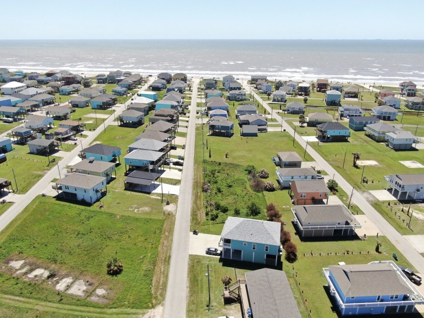 Aerial view of a coastal neighborhood with houses facing the beach and ocean, streets, and green lawns.