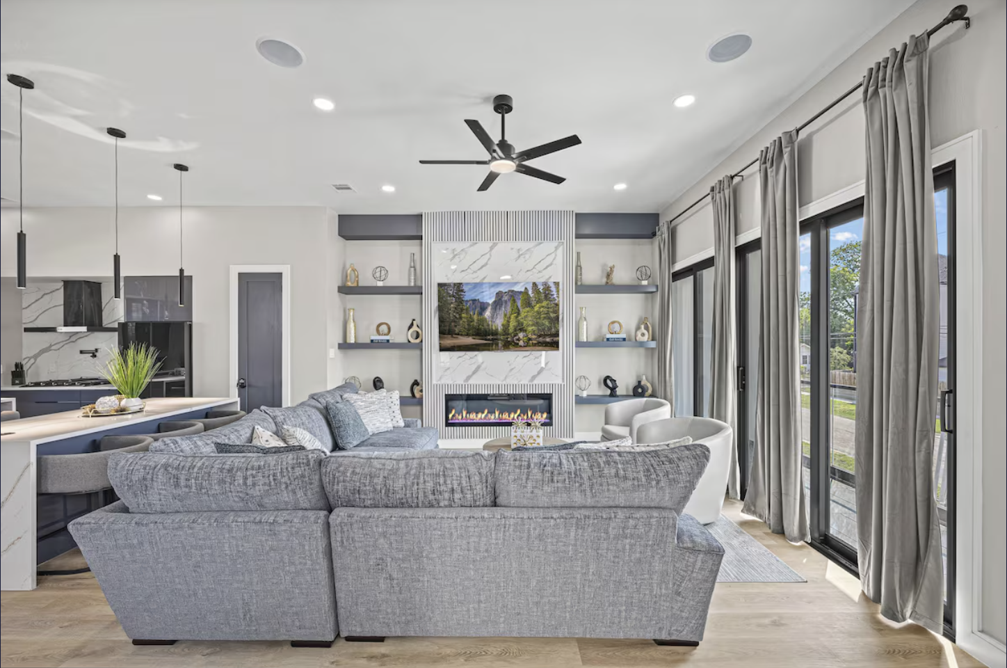 Modern living room with a gray sectional sofa, white and gray armchairs, a fireplace, built-in shelves, a TV, large windows with curtains, and a ceiling fan.