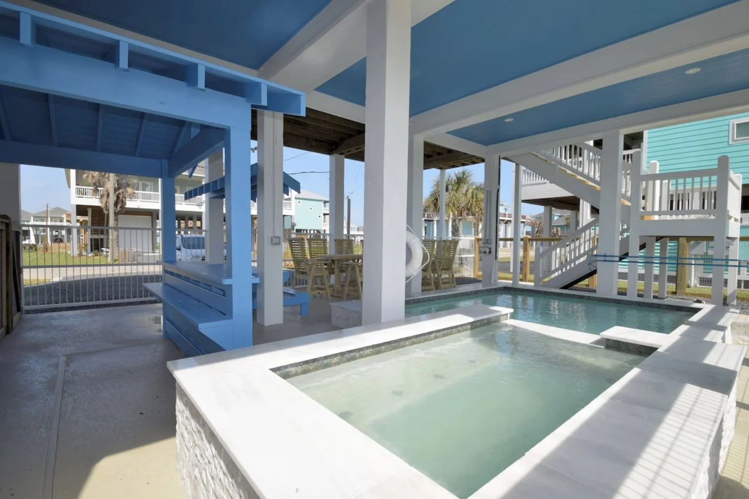 Outdoor pool area with a hot tub, covered seating, and stairs leading up to a second level, surrounded by a white fence and colorful beach houses.
