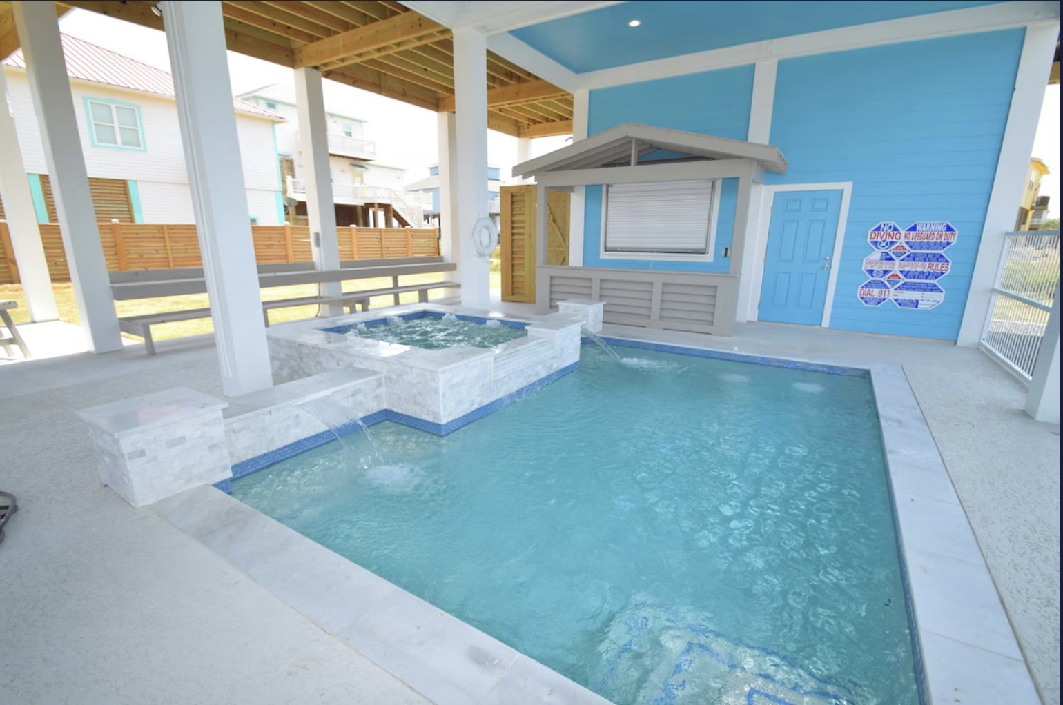 An outdoor pool with a hot tub on one side, under a covered patio area with wooden beams. Blue siding with white trim on a small building features a window with a roller shutter and a nearby door. There are cartoon signs on the wall with swimming-rel