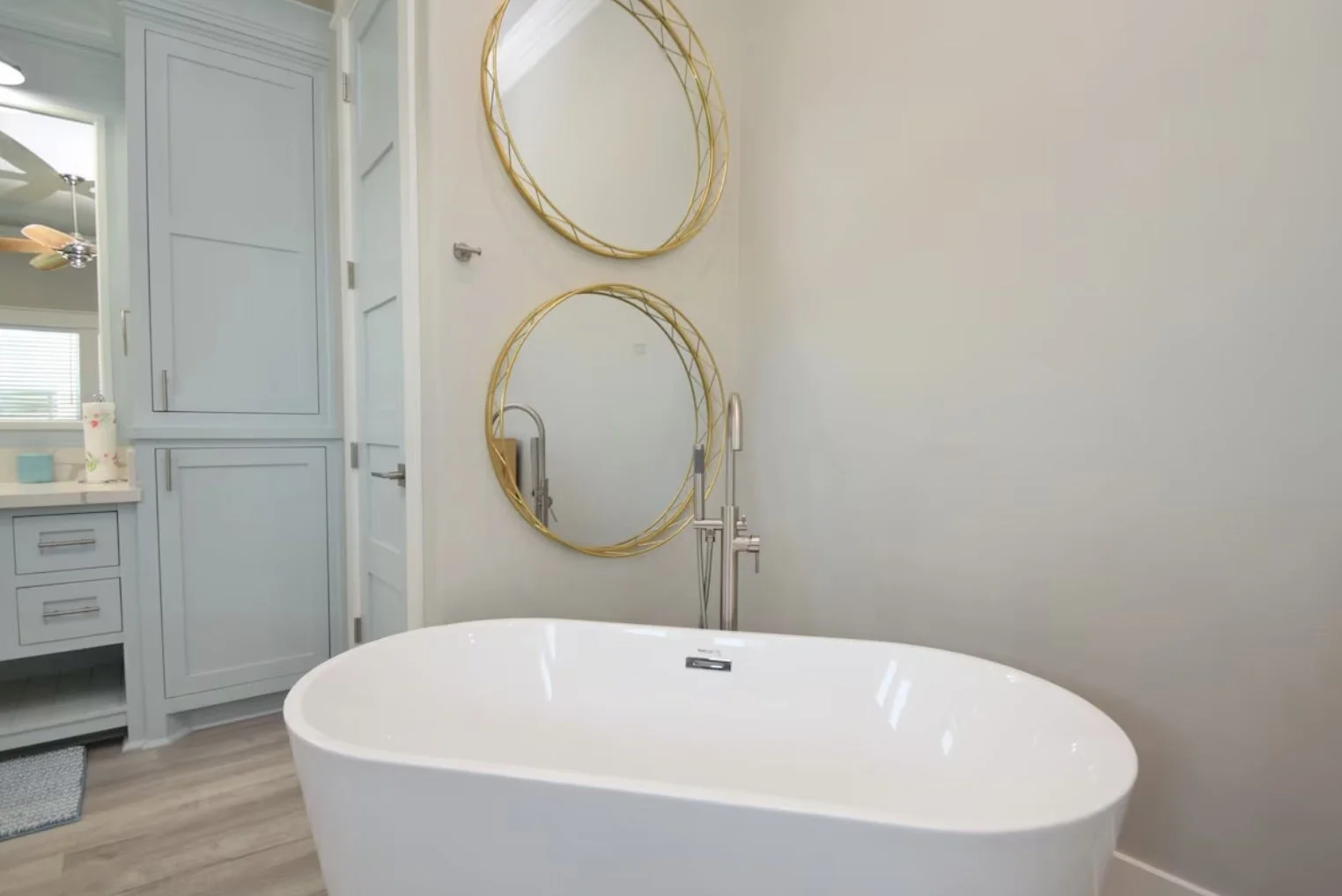 A modern bathroom with a white freestanding bathtub, two circular gold-framed mirrors on the wall, a silver faucet, and a light-colored wooden floor.