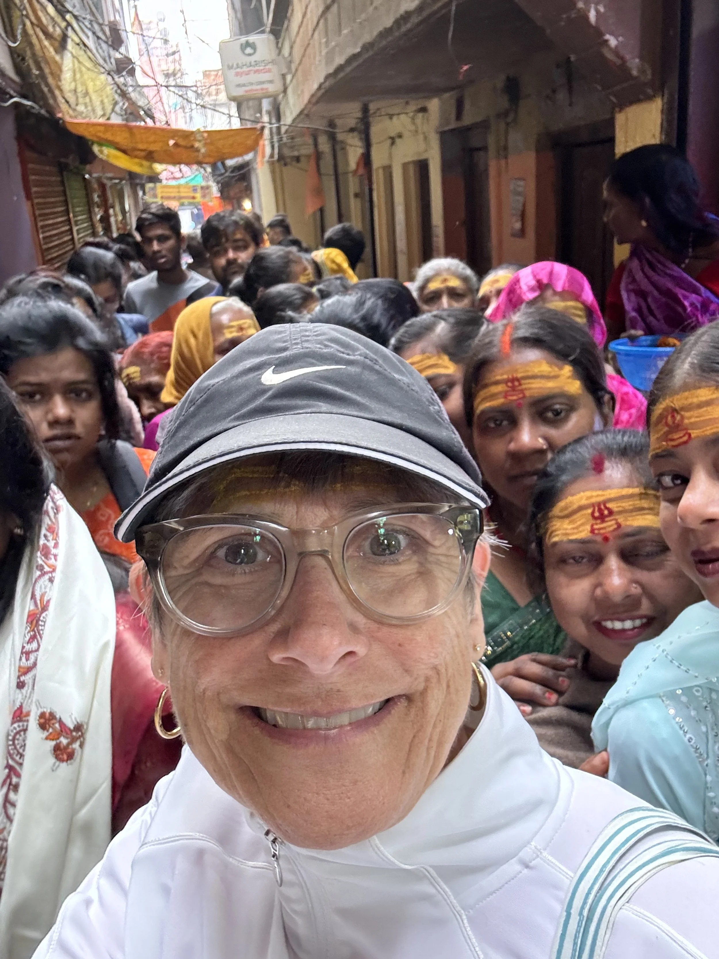 A woman taking a selfie in a crowded street with a large group of women. The women have yellow and red markings on their foreheads and are wearing traditional Indian clothing, with some in colorful scarves and dupattas. The woman in white appears to