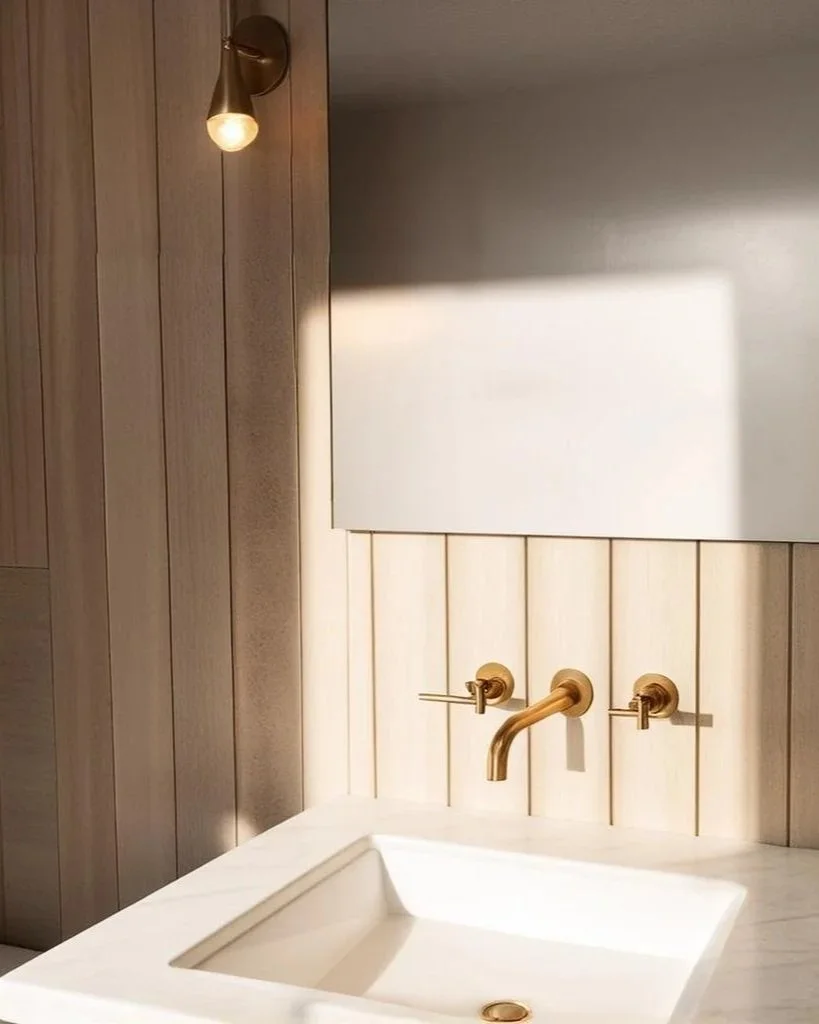 A minimalist bathroom sink with a matte white rectangular basin, paired with a gold wall-mounted faucet and matching handles, set against light vertical wood paneling. A small wall-mounted light fixture is illuminated above.