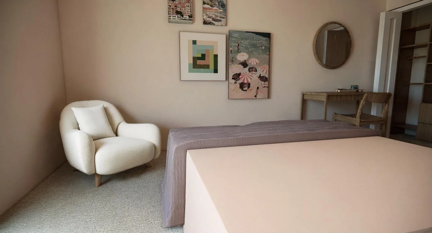 A corner of a bedroom with a cream-colored armchair, a bed with gray bedding, and a small wooden desk with a mirror above it, hanging on a wall decorated with framed artwork.