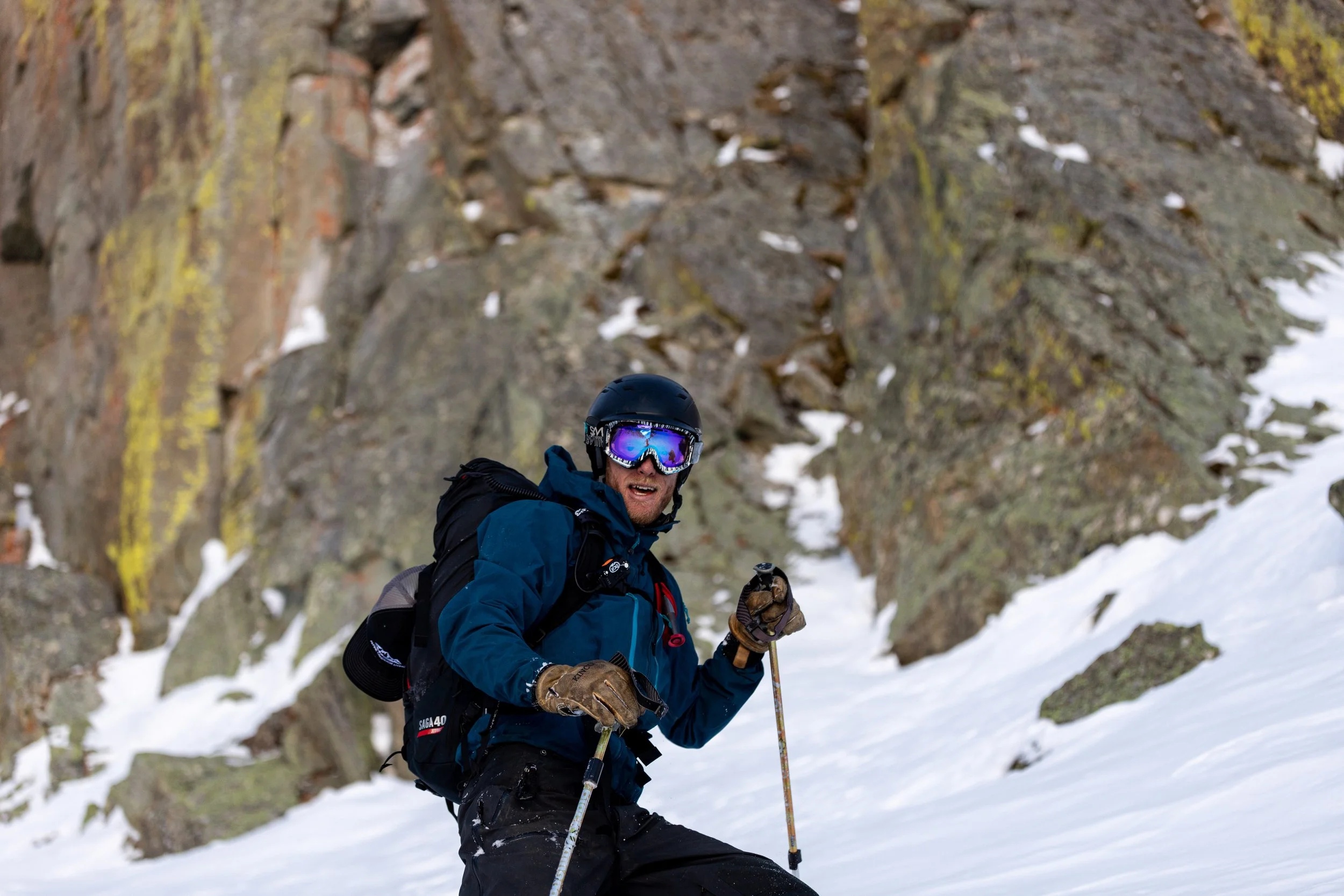 Man in an orange jacket and green pants posing with arms outstretched on a snow-covered street in a small town, with buildings and cars in the background during snowy weather.