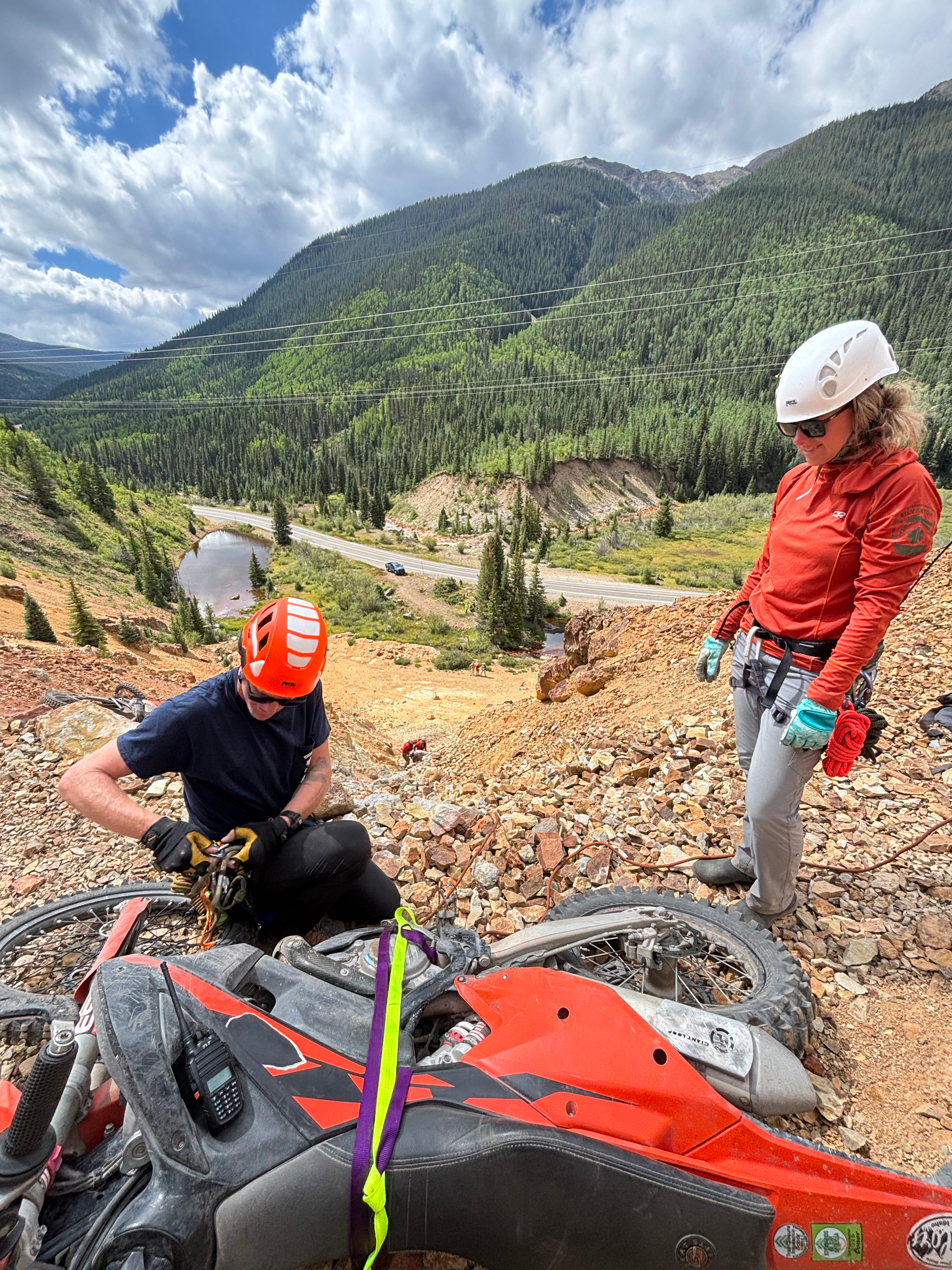 Two people wearing helmets and protective gear repairing a dirt bike on a rocky trail with lush green mountains, trees, and a cloudy sky in the background.