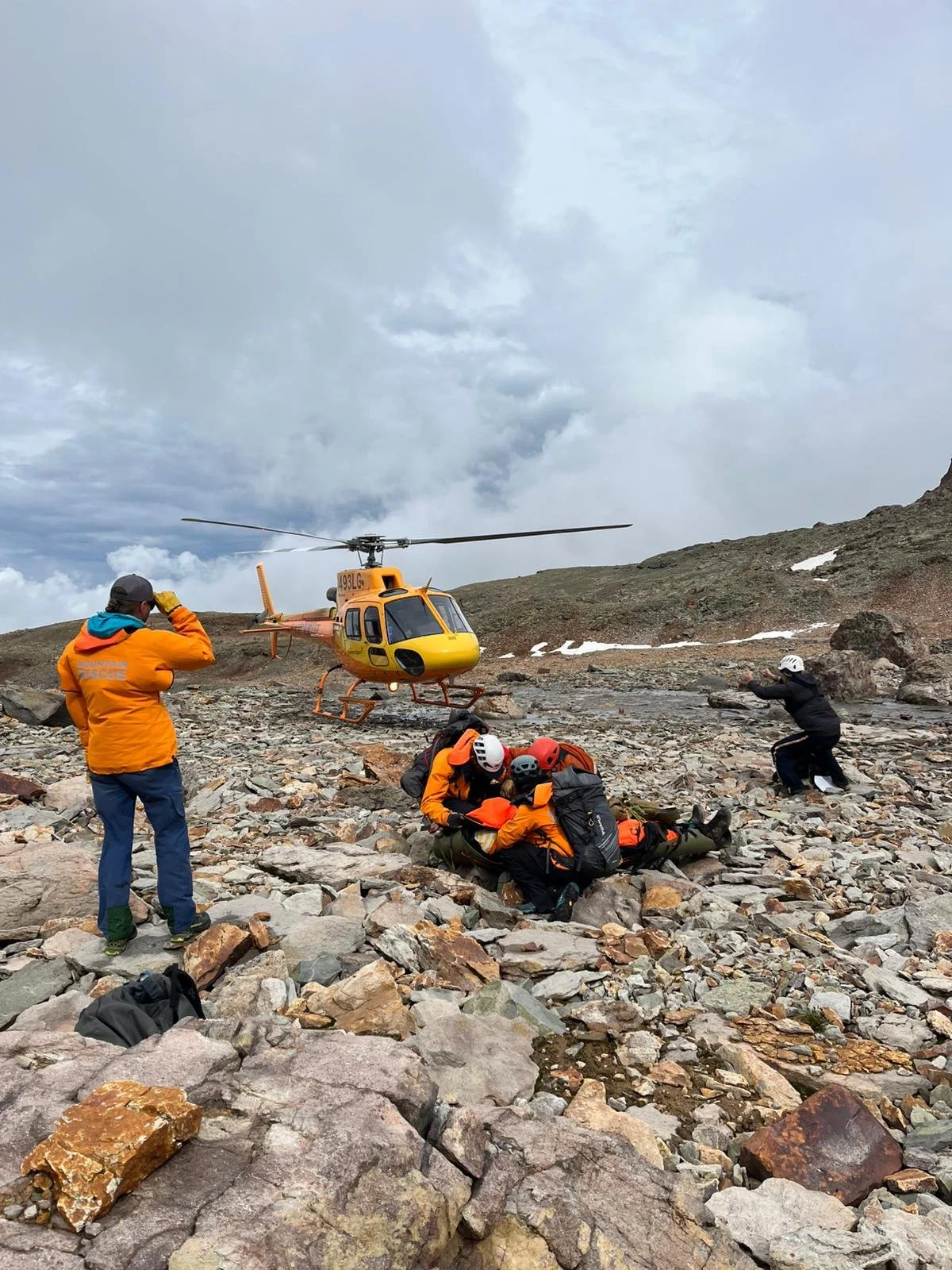 Rescue team assists a person on rocky terrain as a helicopter hovers nearby during a rescue operation in a mountainous area with cloudy skies.