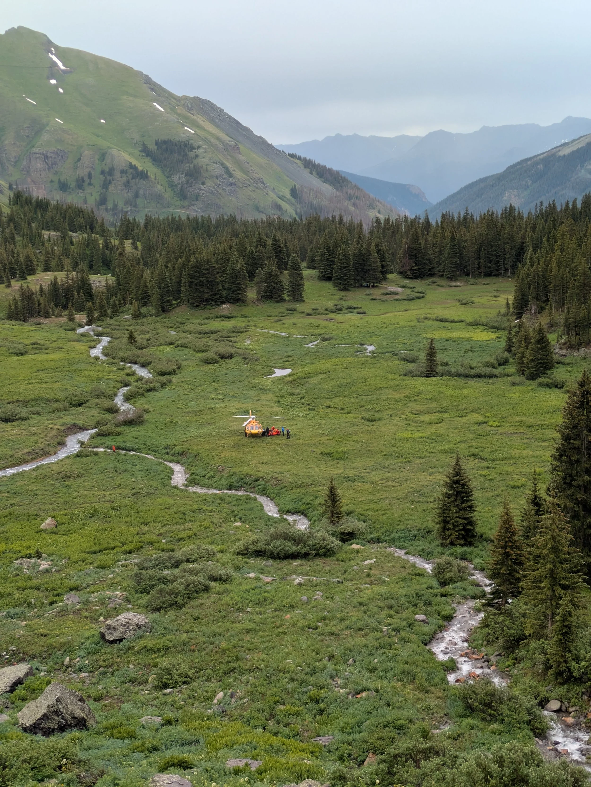 A lush green valley with winding streams, surrounded by dense forests and mountains in the background. A helicopter and equipment are visible near the center of the valley.
