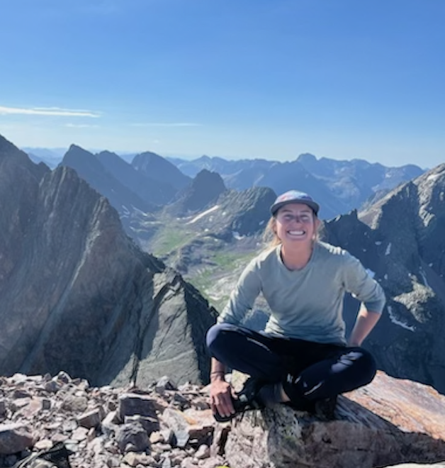 Person sitting on rocky mountain summit with a smile, surrounded by rugged mountain peaks under a clear blue sky.