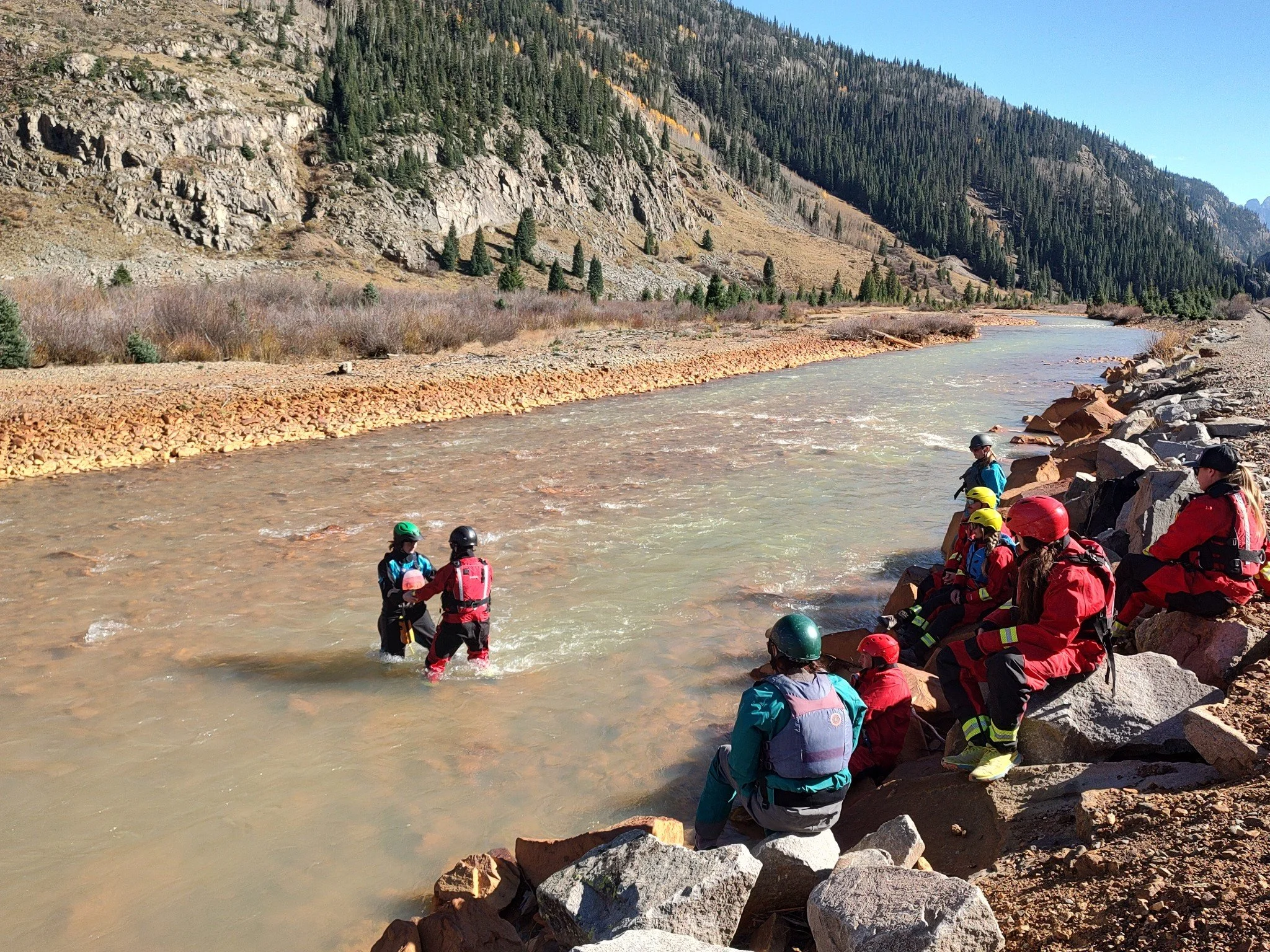 Group of rescue workers wearing helmets and safety gear on rocky riverbank during water rescue training in a mountainous area.