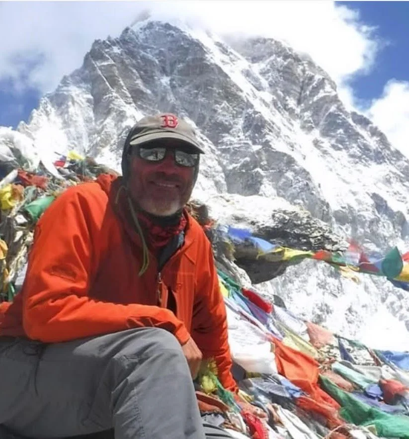 A man in outdoor gear, wearing sunglasses and a cap, sitting on a snowy mountain with prayer flags, with a large snow-covered mountain in the background.