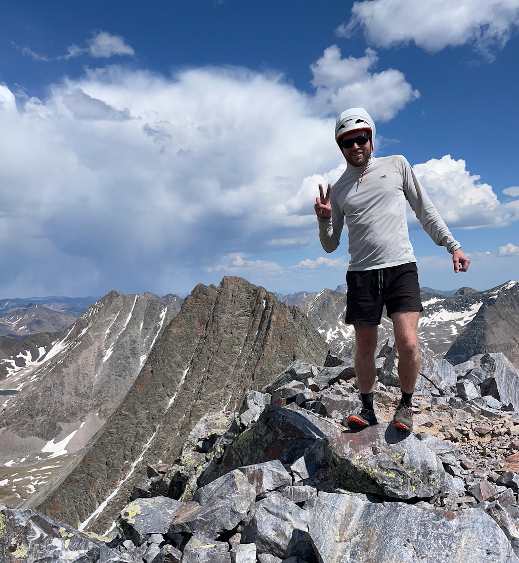 Man wearing a helmet, sunglasses, and athletic gear standing on rocky mountain summit, making a peace sign with his right hand, with scenic mountain range and partly cloudy sky in the background.