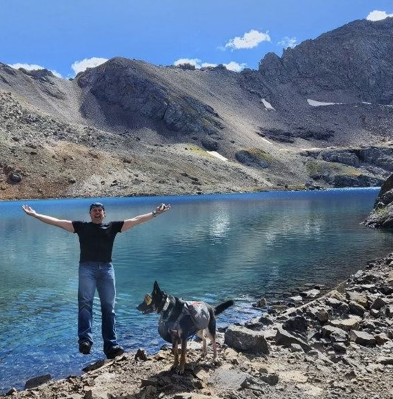 A man stands with arms outstretched and a dog standing nearby on a rocky lakeshore with a mountain range and blue sky in the background.