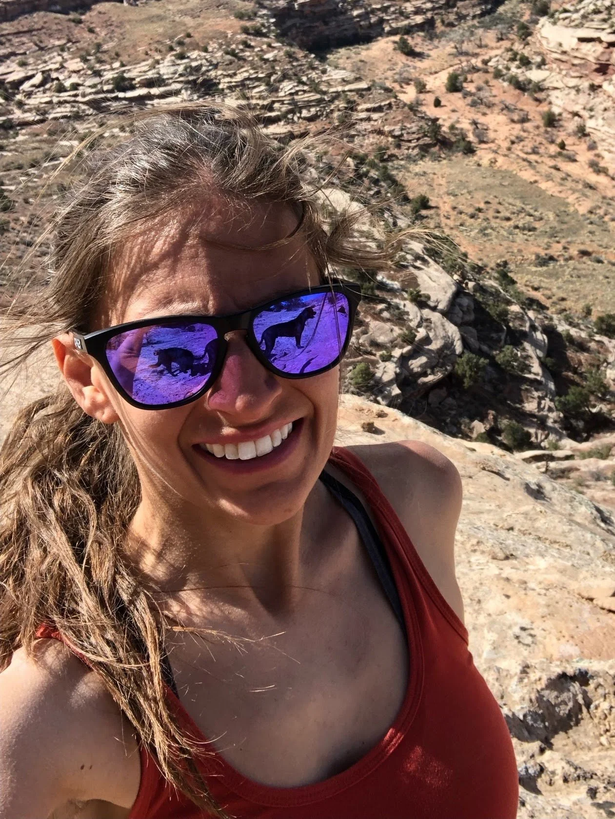 A woman taking a selfie in a rocky, desert-like landscape. She is wearing sunglasses that reflect two bears and a mountain scene, and a red tank top.
