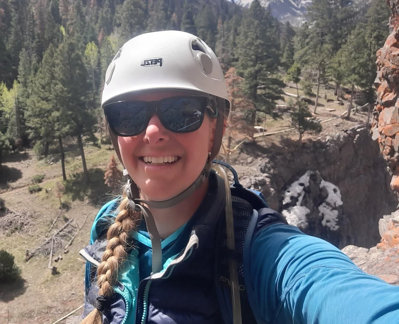 A woman wearing a white bike helmet and sunglasses, smiling and taking a selfie outdoors in a forested mountain area.