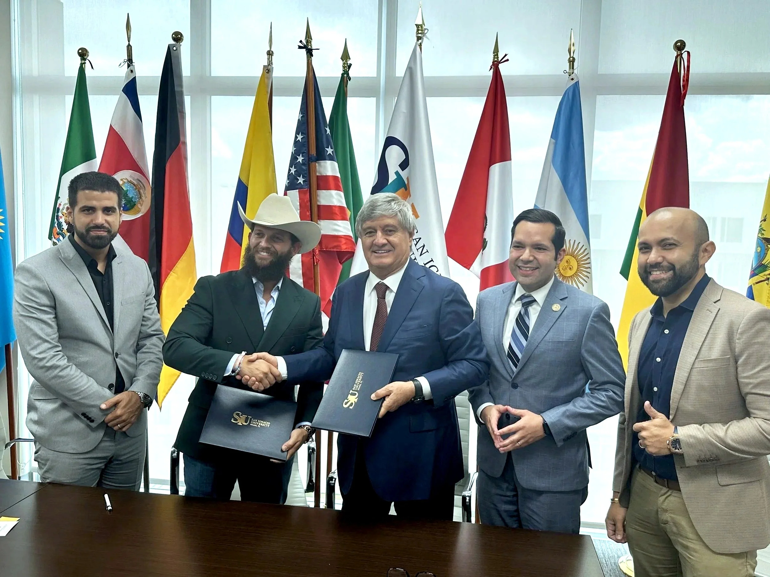 Six men in suits standing at a table, two of them shaking hands, with a background of multiple country flags, in a formal setting.