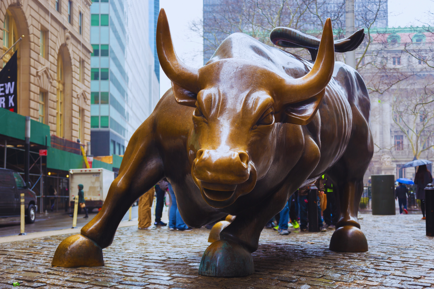 Large bronze statue of a charging bull on a cobblestone street in an urban area with tall buildings and people around.