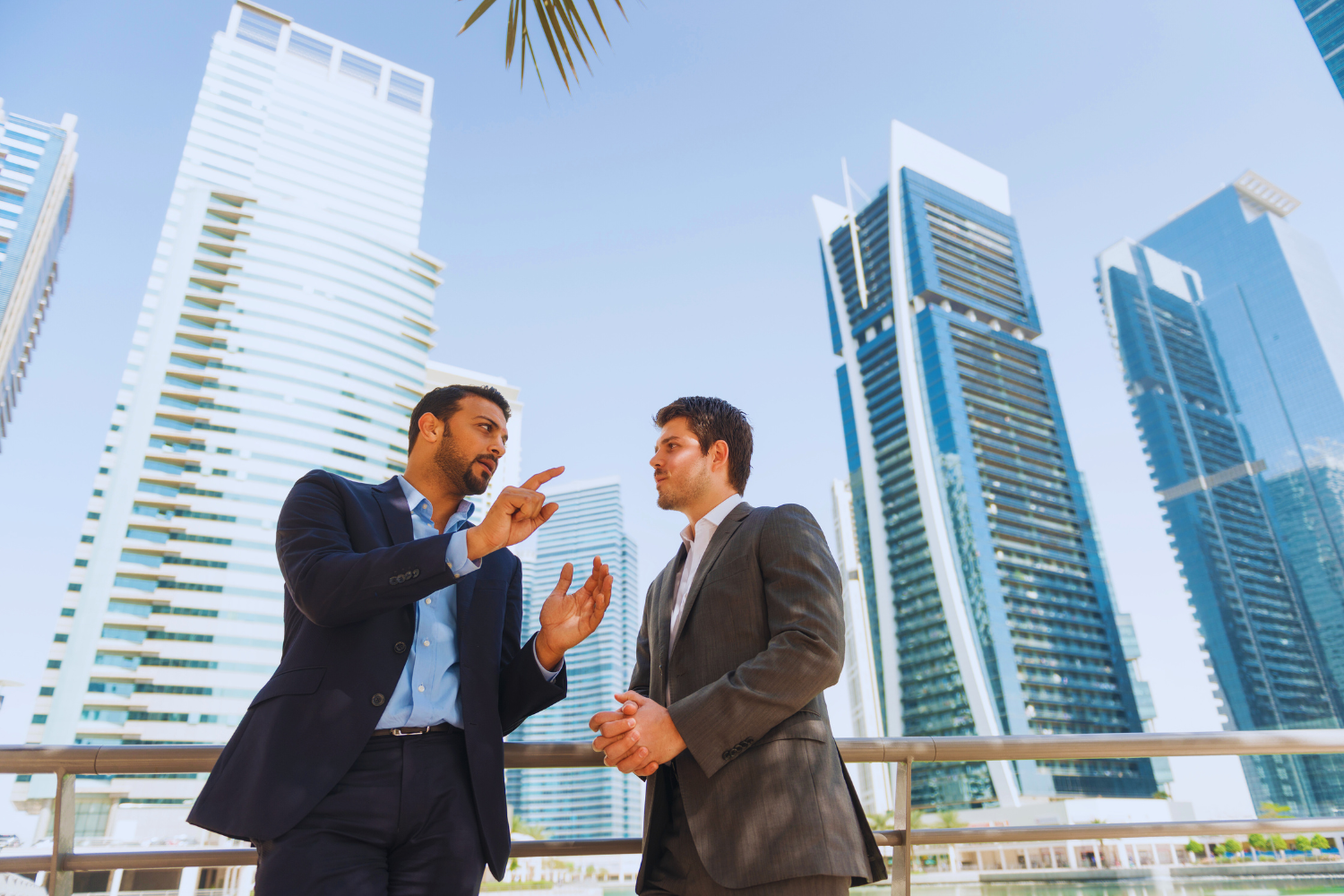Two businessmen in suits having a serious conversation outside in front of tall modern office buildings.