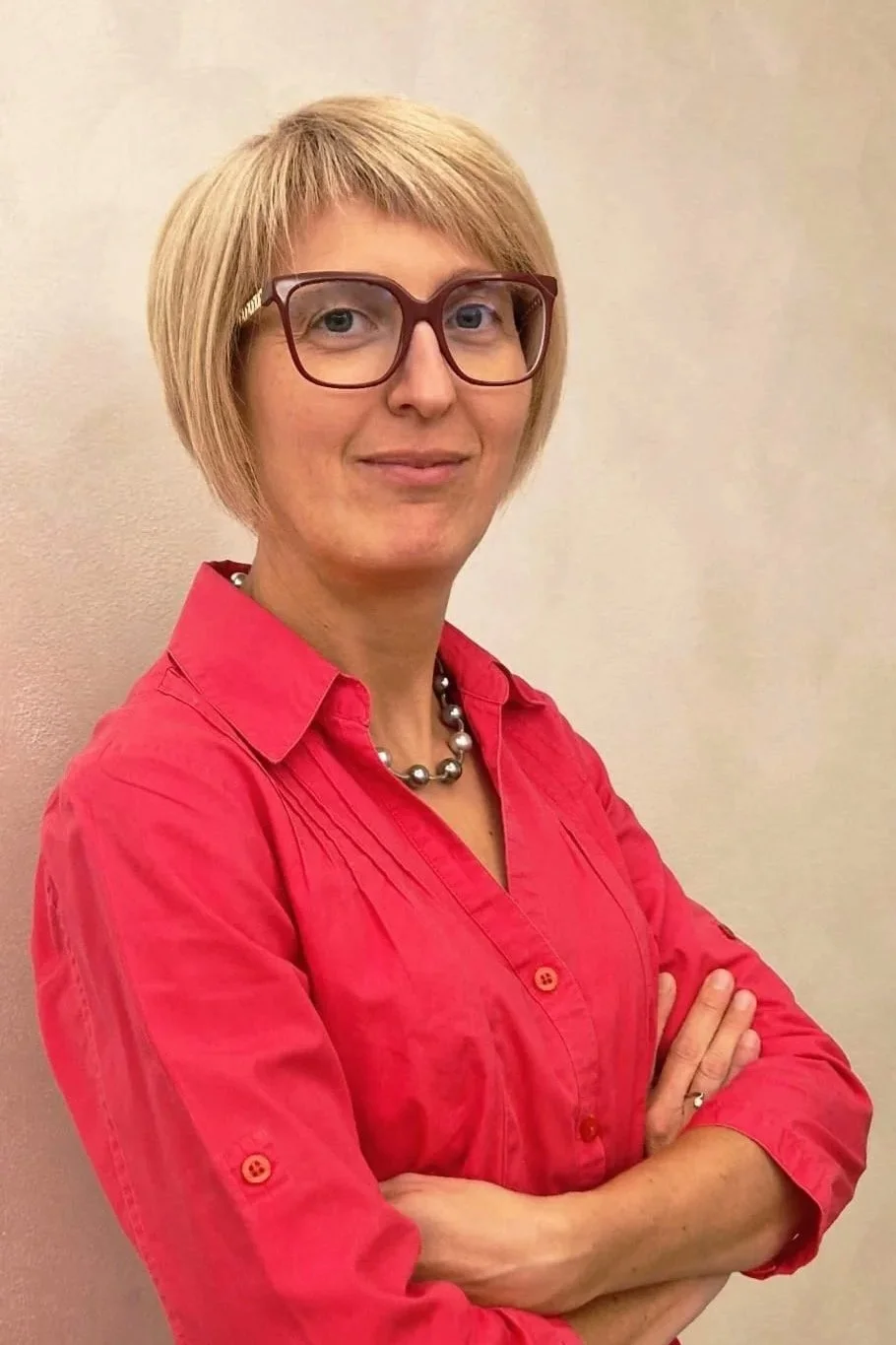 Donna con capelli corti, indossa occhiali con montatura rossa, camicia rossa e collana di perle, si trova contro una parete beige.
