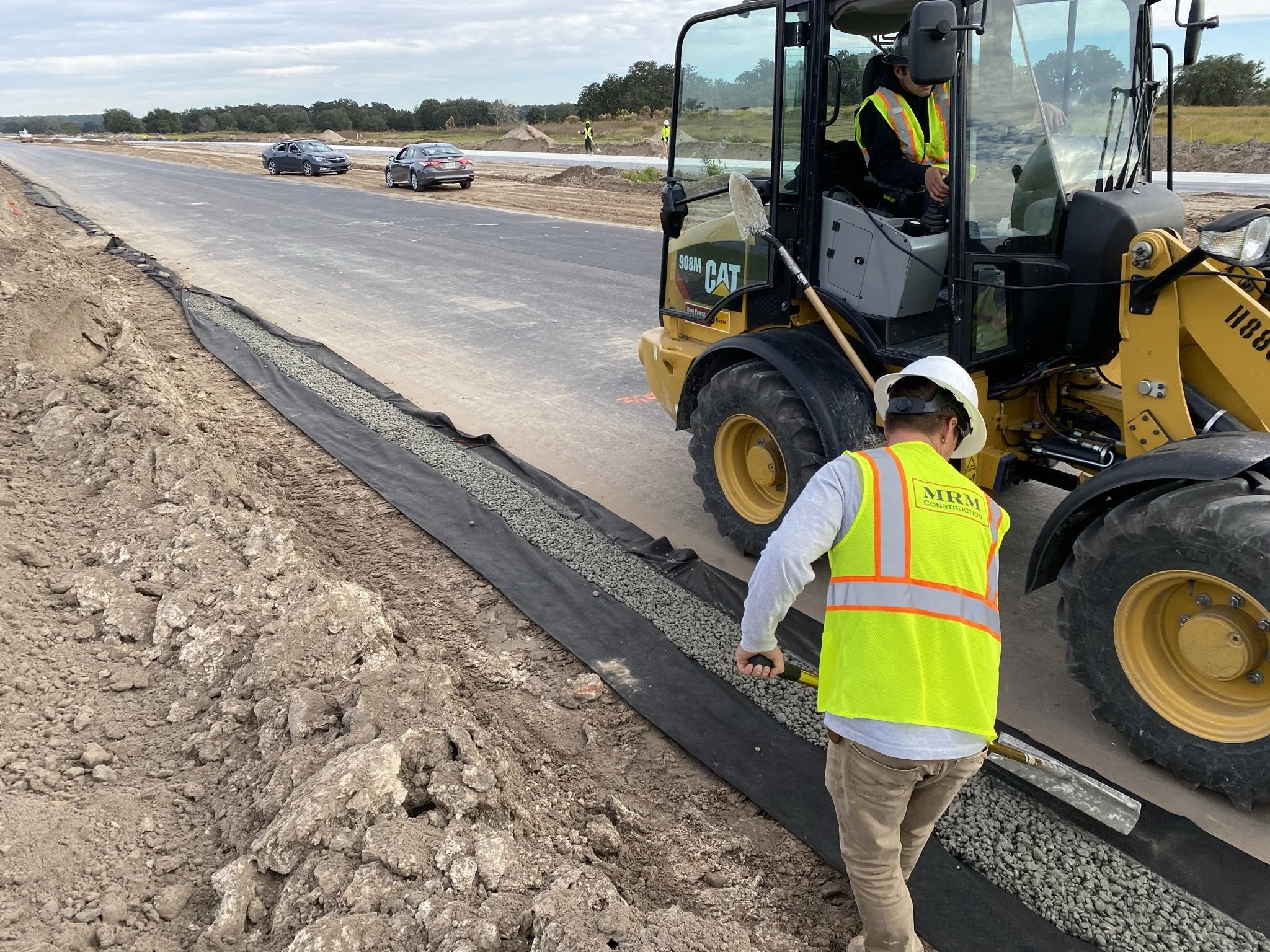 Construction worker with high-visibility vest and helmet using a tool on a newly paved road with a worker operating a small construction vehicle nearby. Several cars are driving on the road, and construction materials are visible along the roadside.