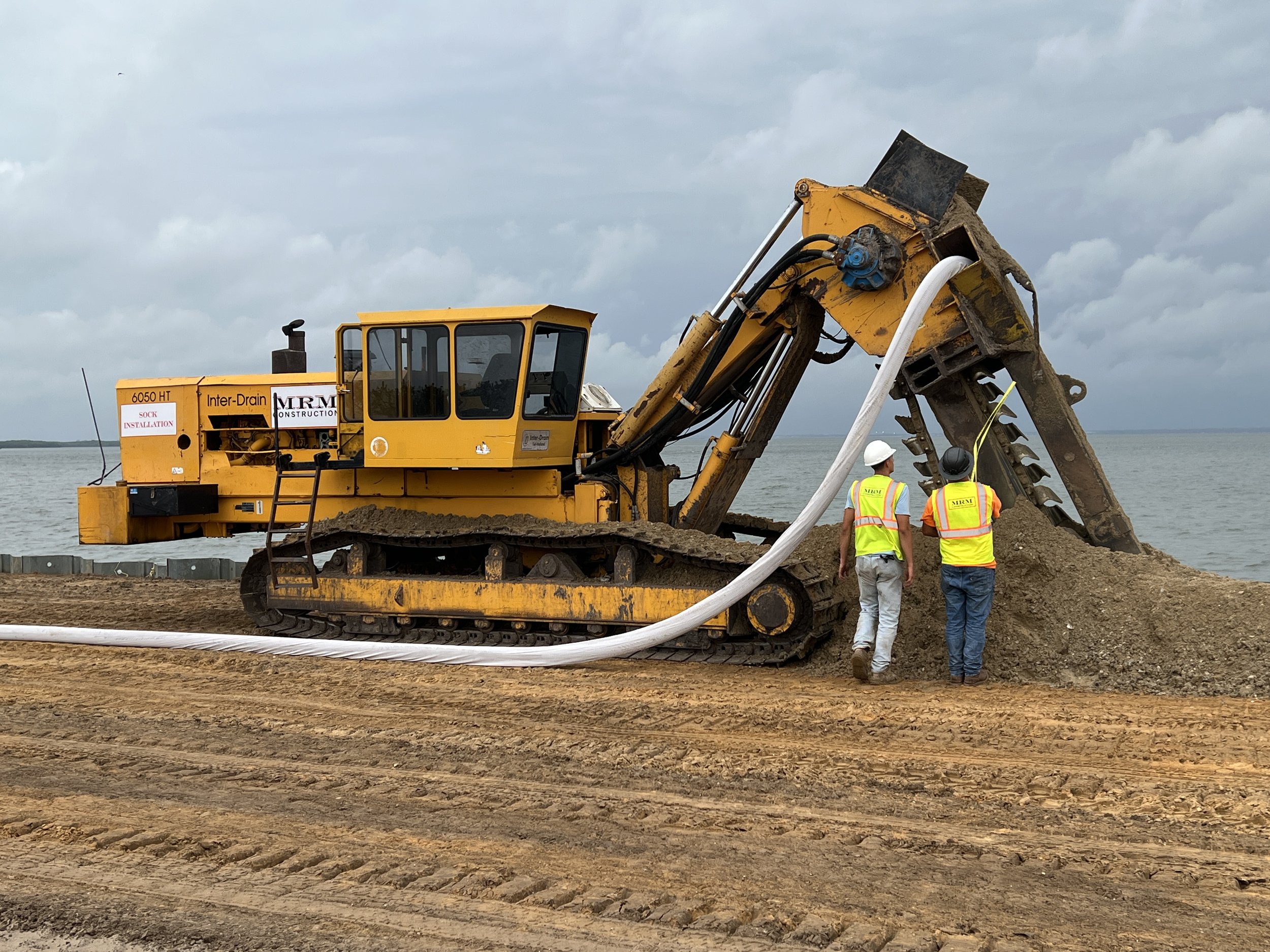 Construction workers in safety vests and helmets working near a large yellow excavator on a dirt construction site by water.