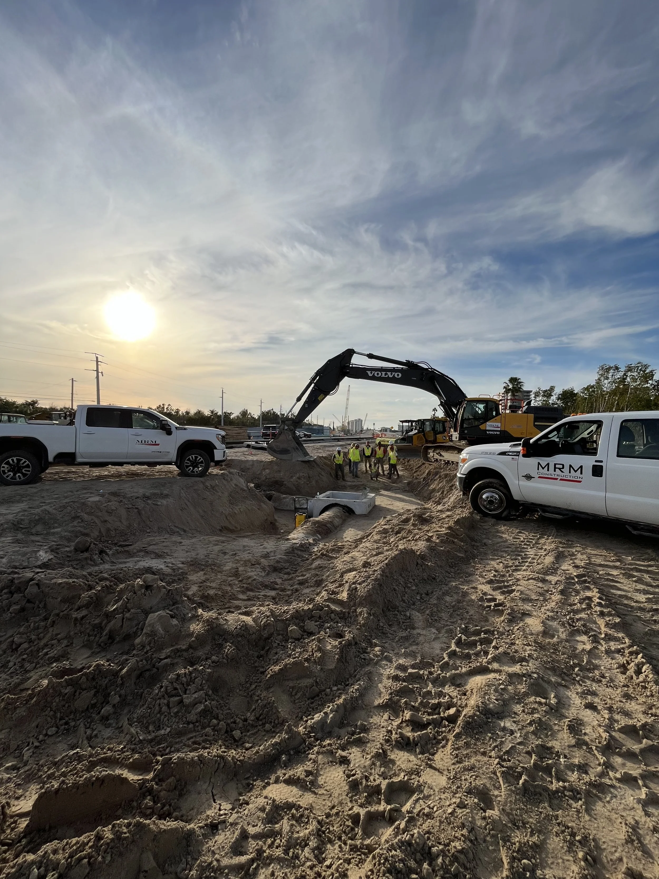 Construction site with an excavator digging in the dirt, workers wearing safety vests in the background, and trucks parked nearby during sunset.