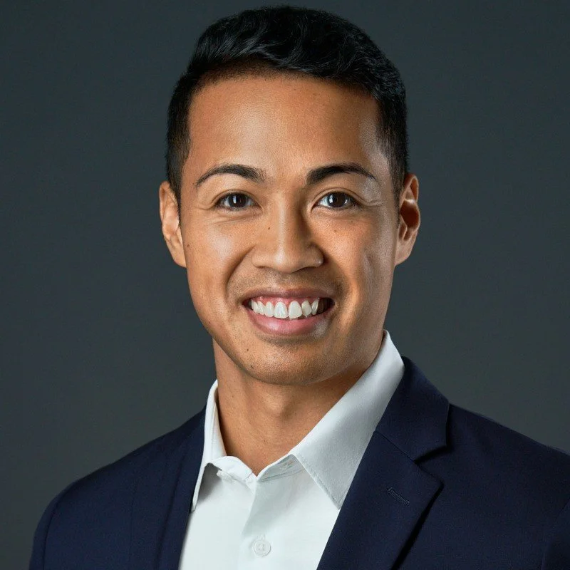 A young man with short black hair, wearing a white collared shirt and dark blazer, smiling against a gray background.