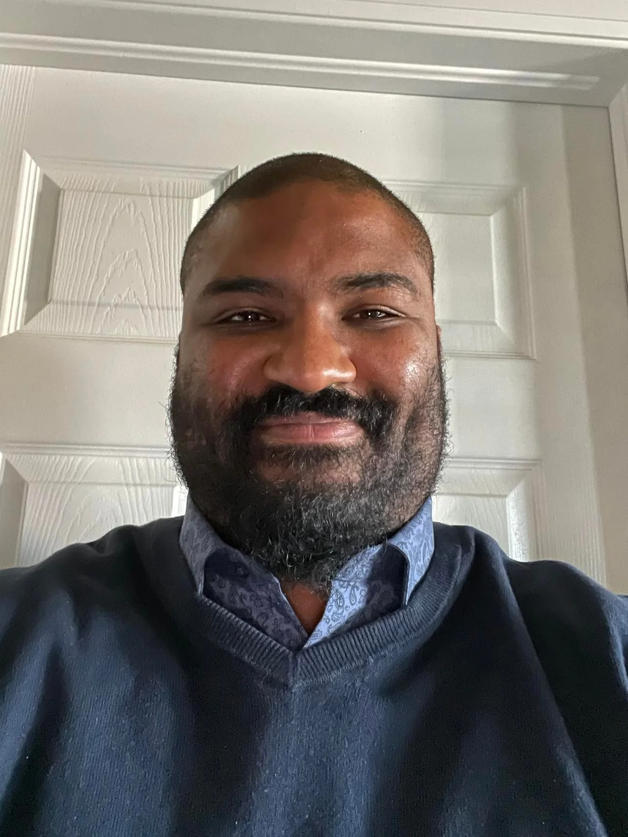A man with a beard smiling, wearing a blue collared shirt and a dark sweater, standing in front of a white door.