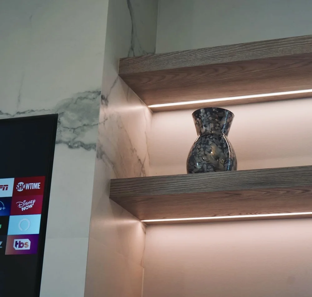 Angled detail of woodgrain shelf edges and linear lighting beside a marble-look television surround