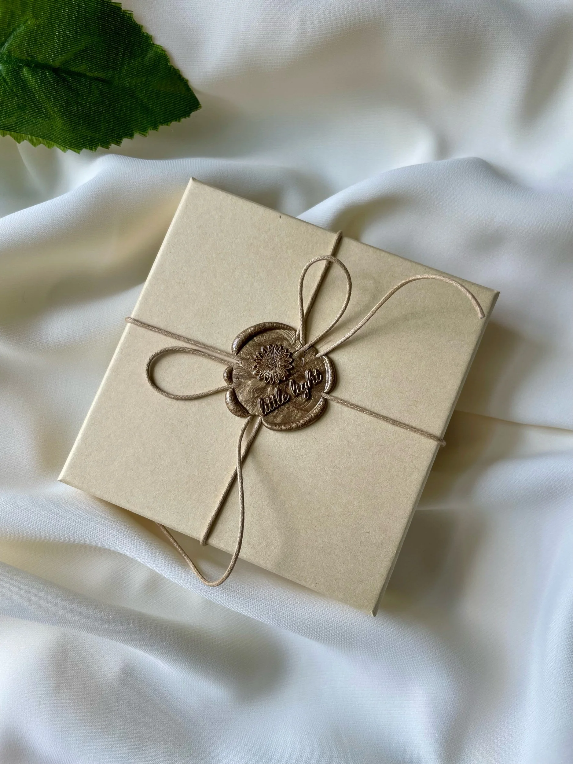 A beige gift box wrapped with a beige string and a wax seal with floral design and the words 'little light' on it, placed on white fabric with green leaves partially visible in the background.