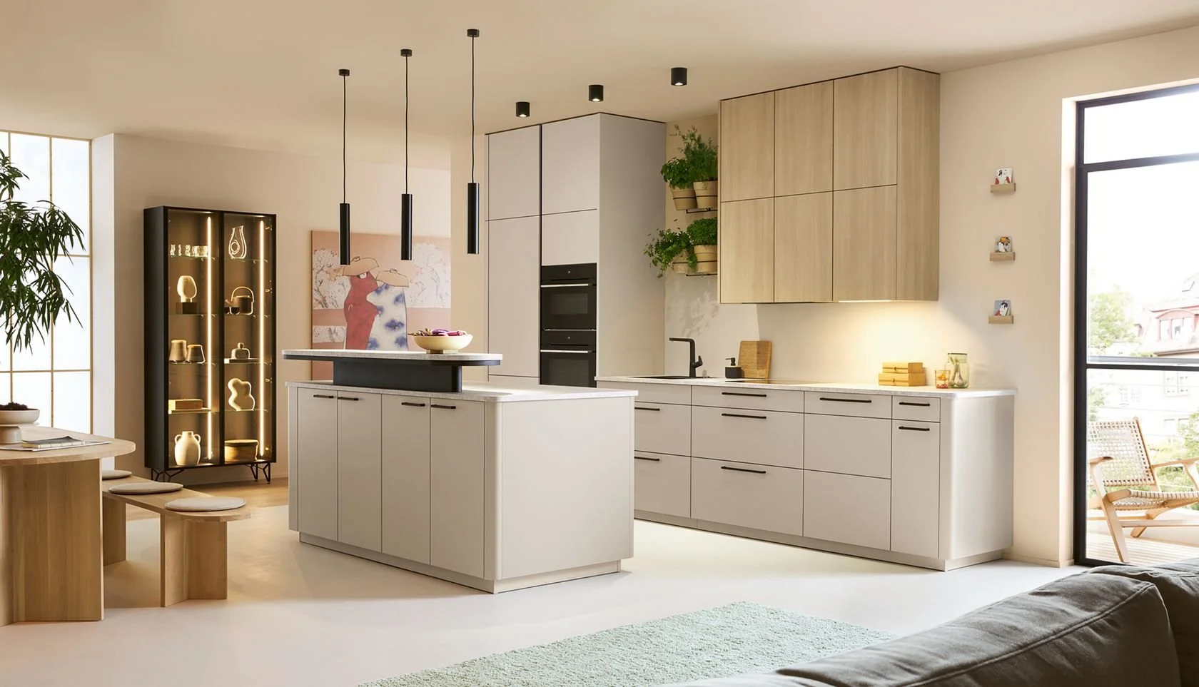 Modern kitchen with white cabinets, a kitchen island, black handles, a sink, built-in oven, and wooden wall cabinets. There's a large window and a glass door leading to an outdoor balcony with seating. Decor includes shelves with plants and small decorative items.