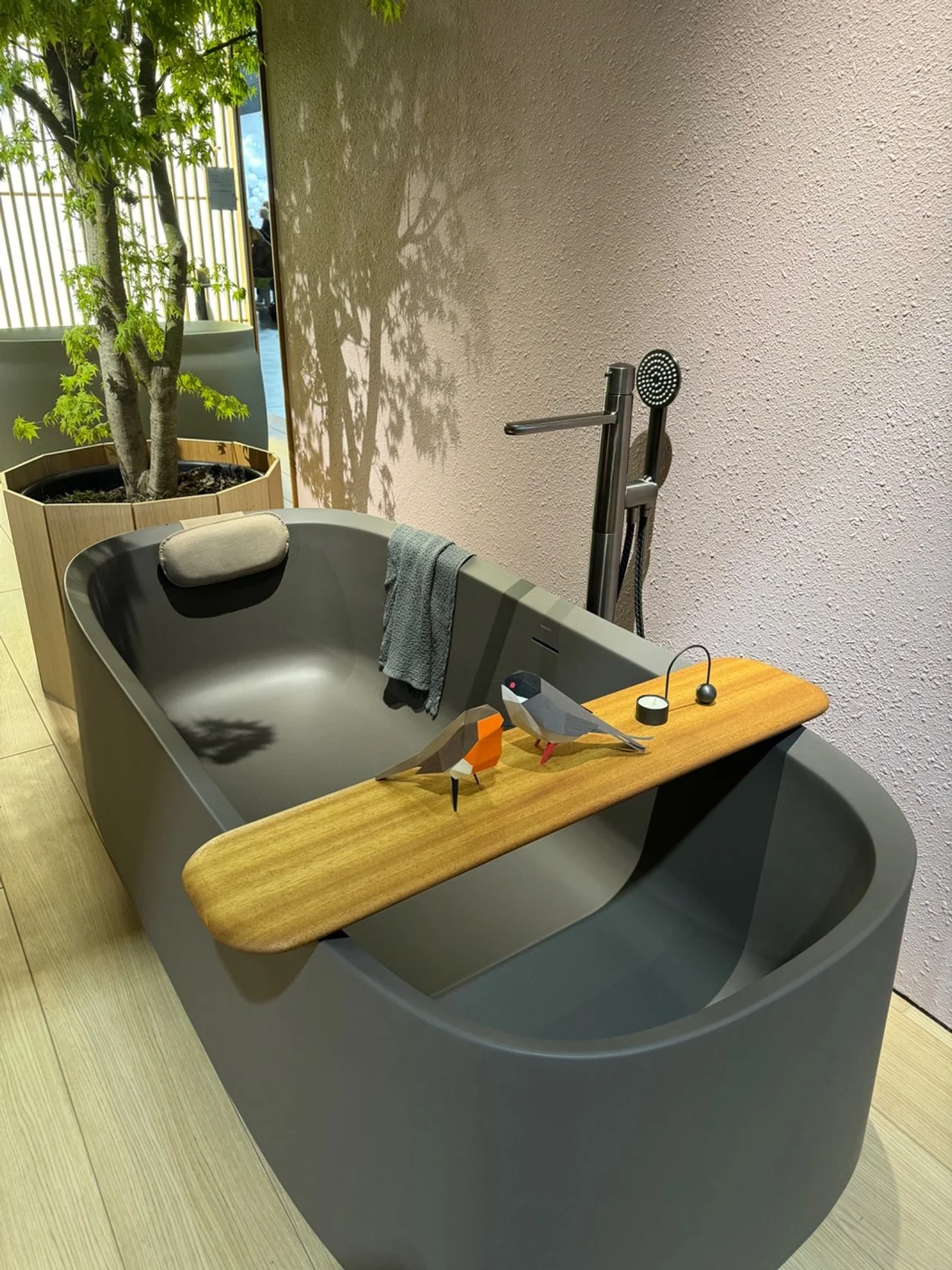 Modern gray freestanding bathtub with a wooden tray holding two origami-style bird figures, a small black and white container, and a black spherical object. Towel draped over the bathtub edge. A shower handheld massager near the wall. A potted plant with green leaves casting a shadow on the wall.