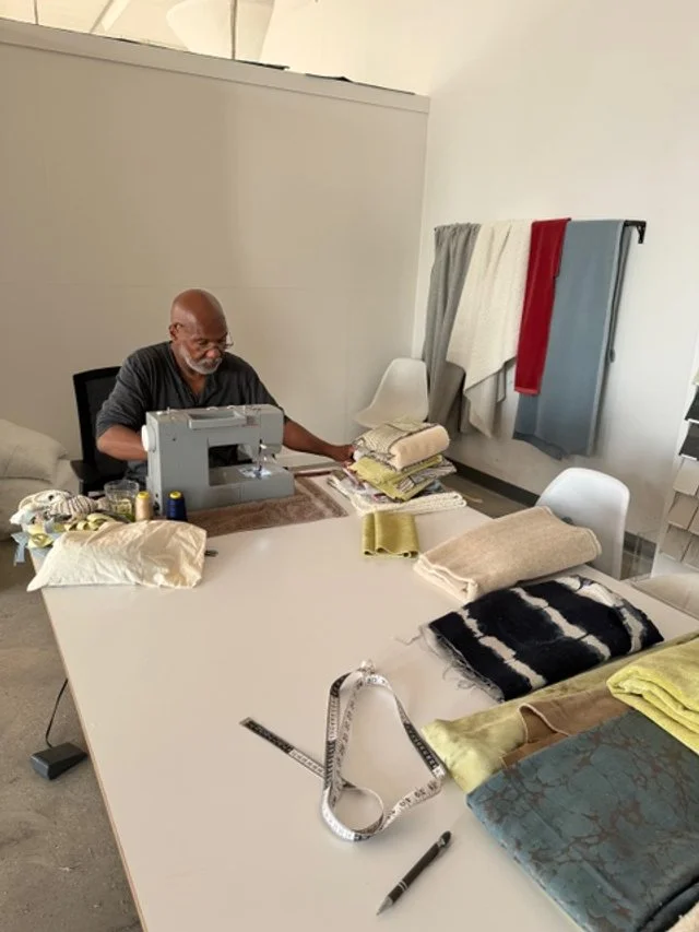 A man sewing at a table surrounded by fabric swatches and sewing tools, with towels hanging on a rack in the background.