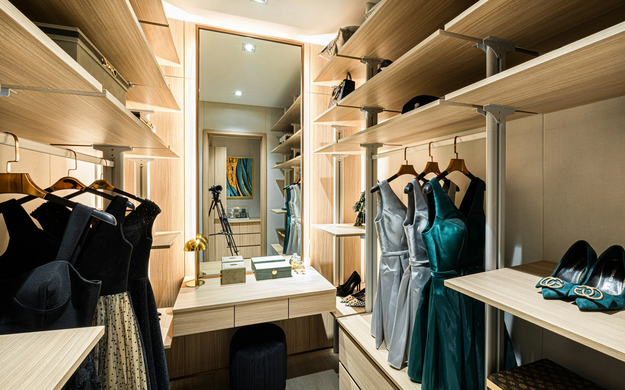A walk-in closet with shelves and hanging space for clothes, including black, silver, and green dresses, a pair of teal shoes, and a mirror, with a small desk and a camera on a tripod.