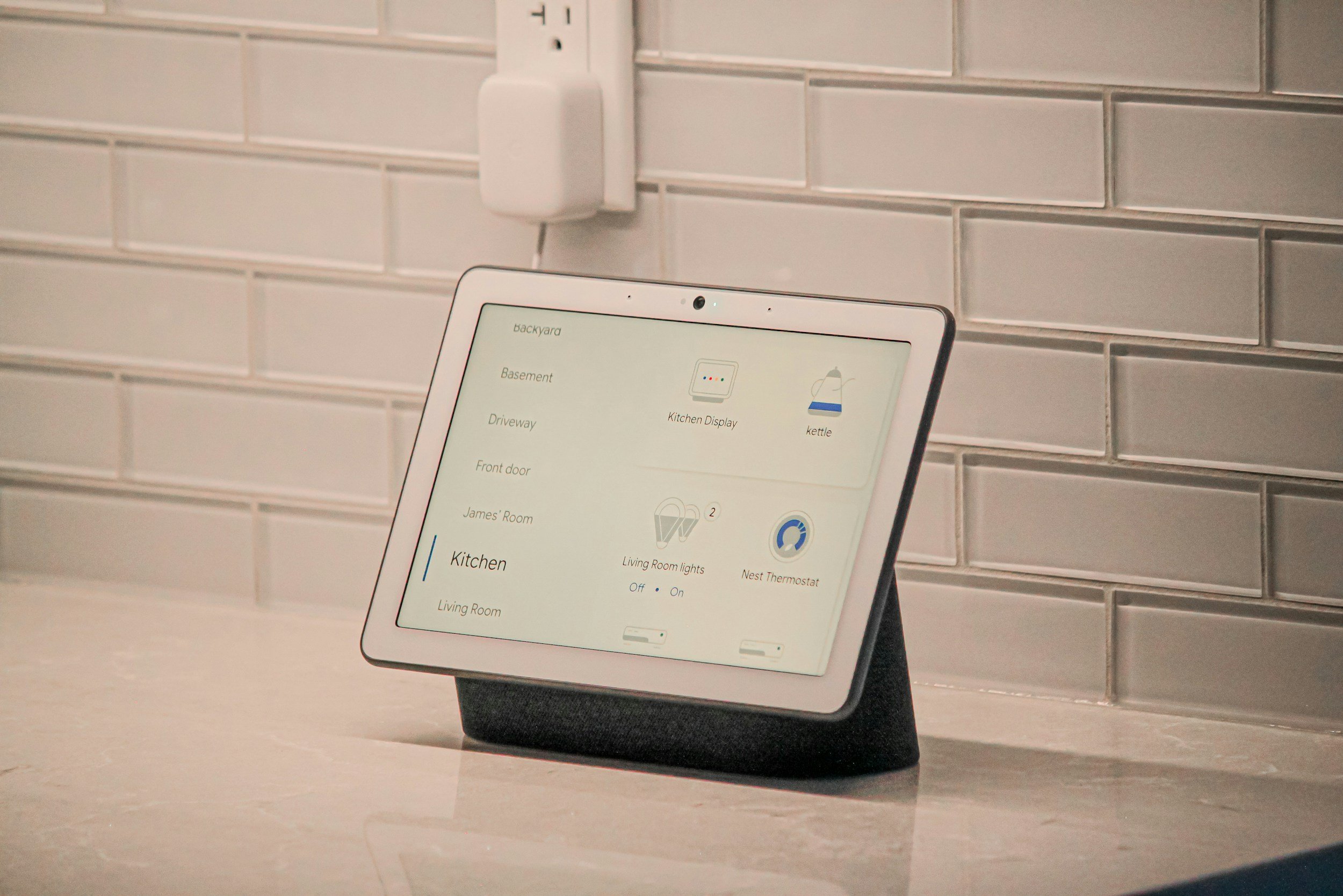 A smart home control device on a countertop, displaying various room options and device statuses, with a gray subway tile backsplash and a wall outlet in the background.
