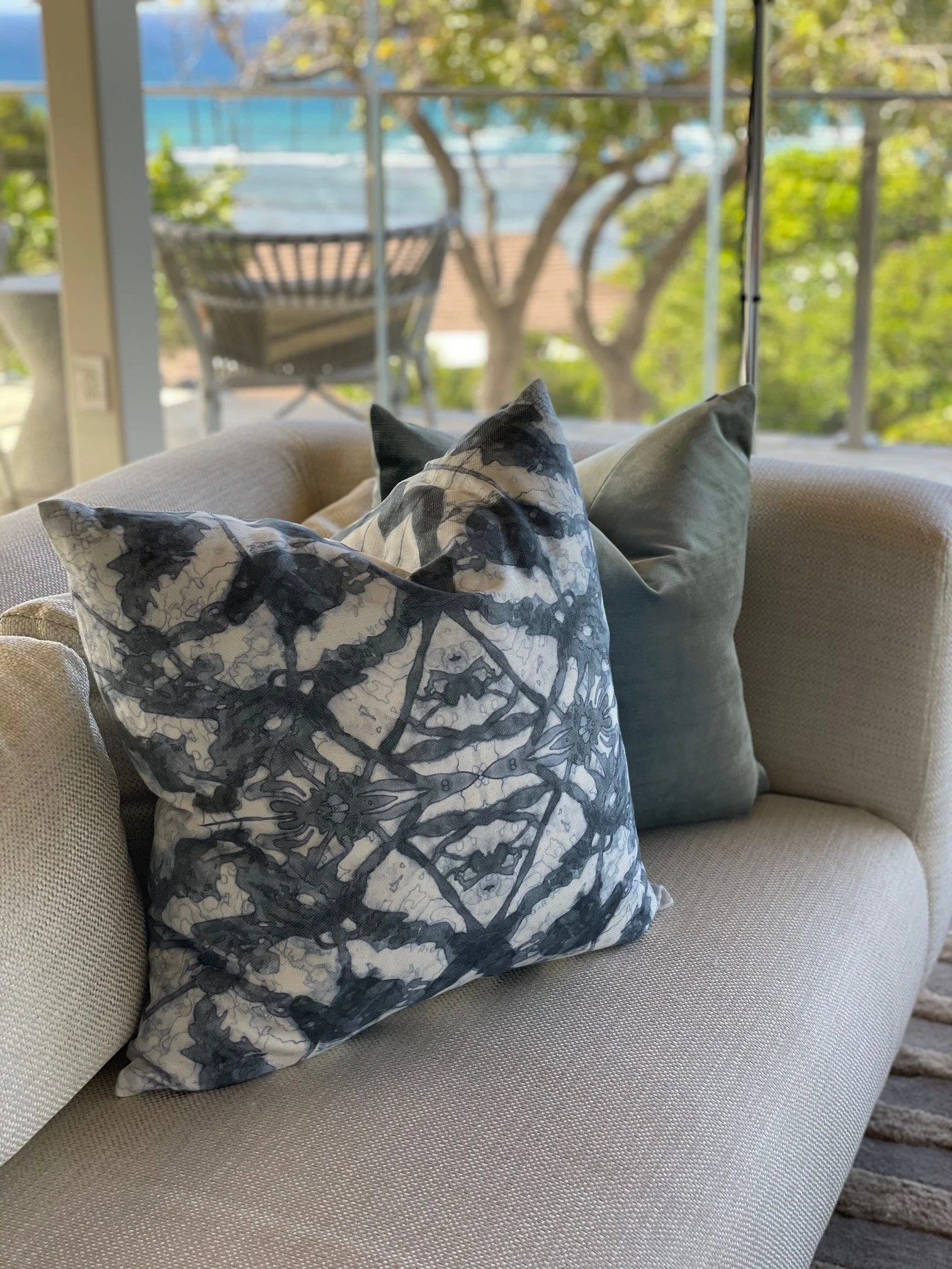 A beige sofa with three decorative pillows: one with a black and white leafy pattern, the second a solid dark green, and the third a solid light beige. Behind the sofa, a large window shows a view of trees and a balcony with outdoor furniture.