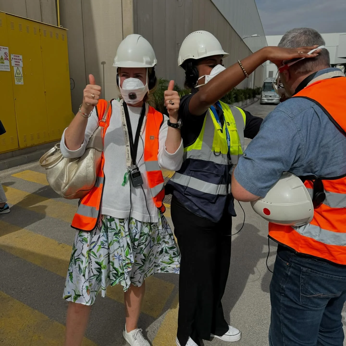 Group of people wearing safety vests, helmets, and face masks outdoors near a building, with one person giving a thumbs-up and another adjusting their safety helmet.