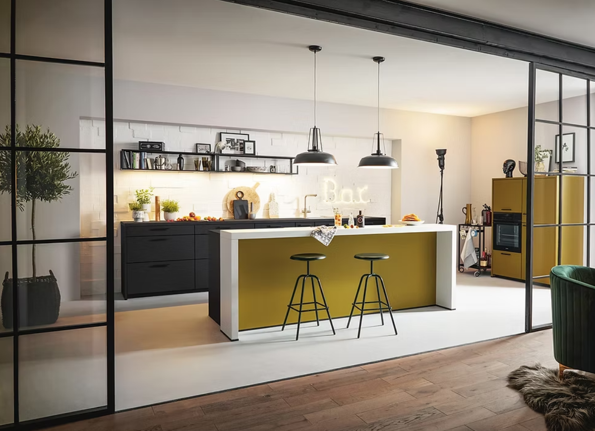 Modern kitchen with black cabinets, an island with olive green base and white countertop, two black barstools, yellow storage cabinet, and decorative elements, viewed through a glass partition.
