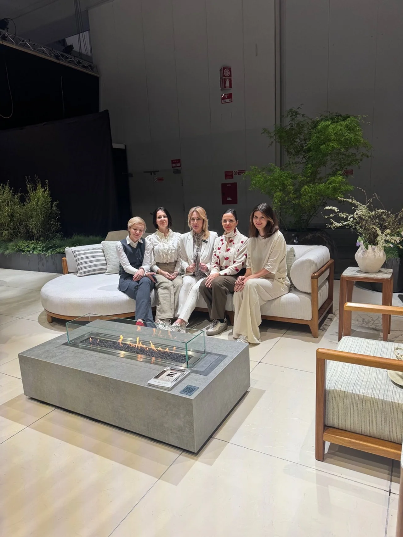 Five women sitting on a white sofa in a modern indoor space with a fire feature in front of them, greenery, and minimalist furniture.