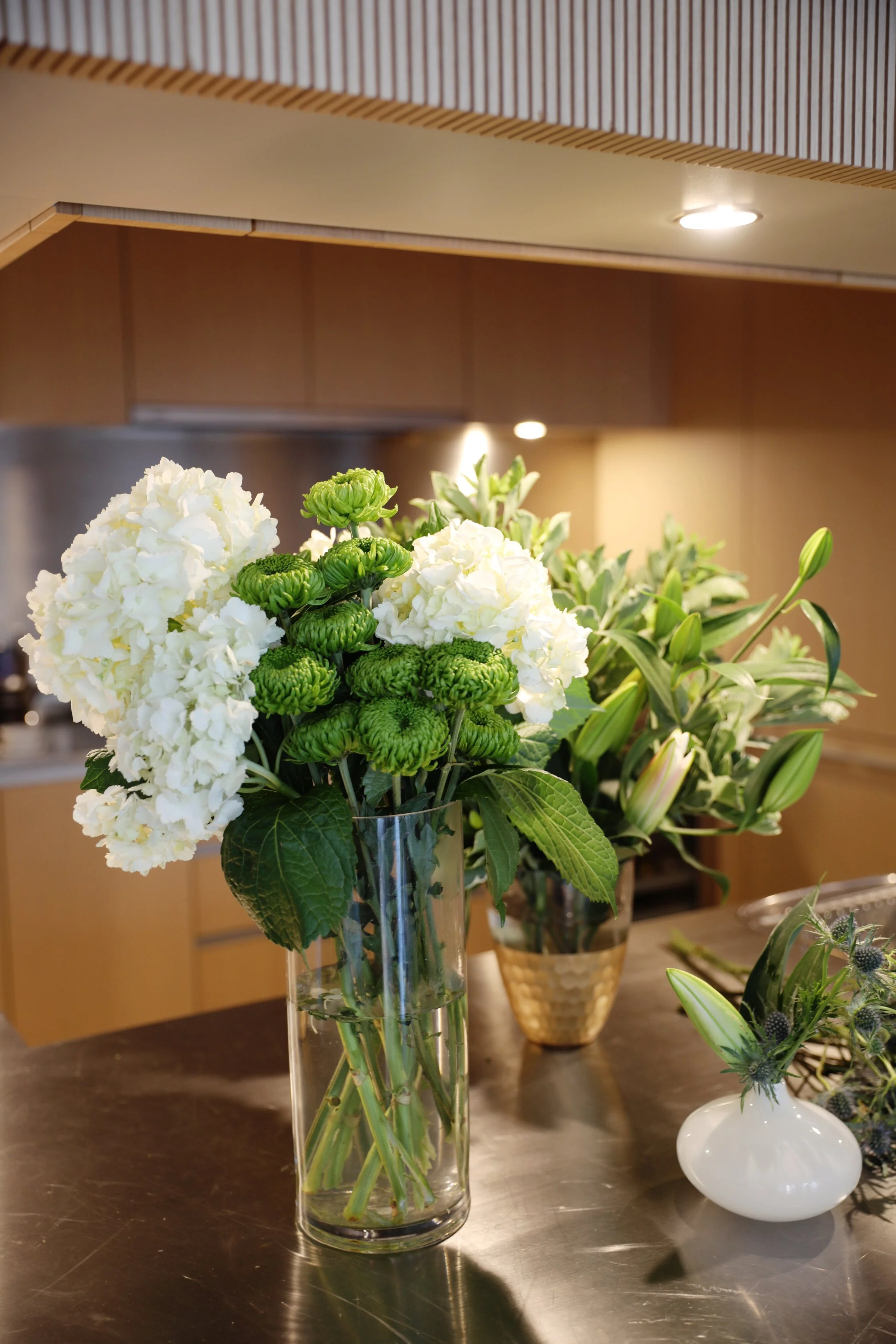A glass vase with white hydrangeas, green chrysanthemums, and leaves on a kitchen table. In the background, there is a wooden kitchen wall with some vase with pink lilies and another white vase with purple thistles.