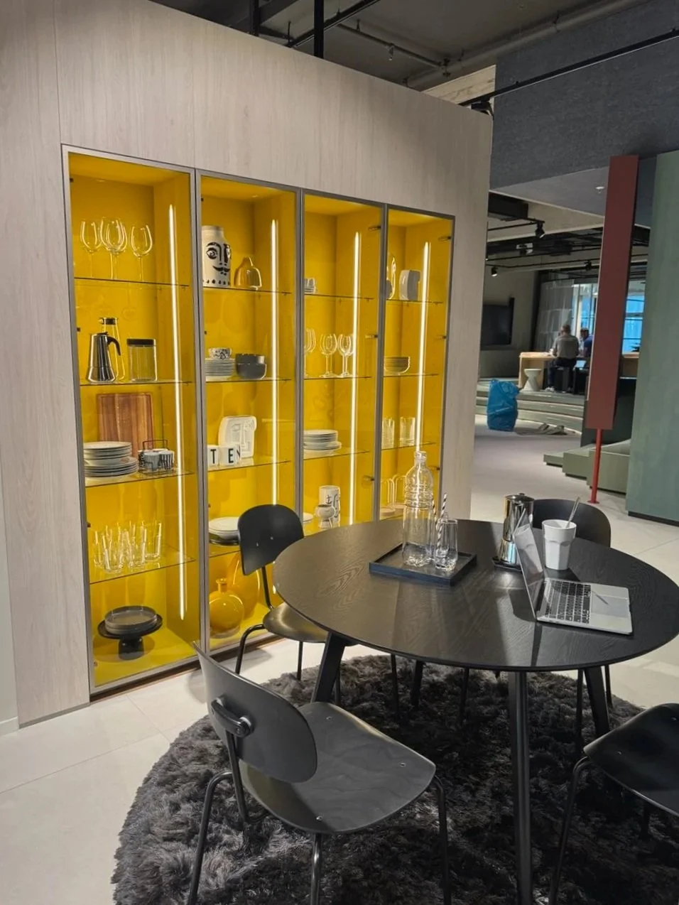 A modern meeting or dining area with a black round table, three black chairs, and a dark gray shaggy rug. In the background, there's a yellow display cabinet with glass doors showcasing glassware, dishes, and decorative items. The room has a contemporary industrial design with exposed ceiling pipes and a mix of light and dark colors.