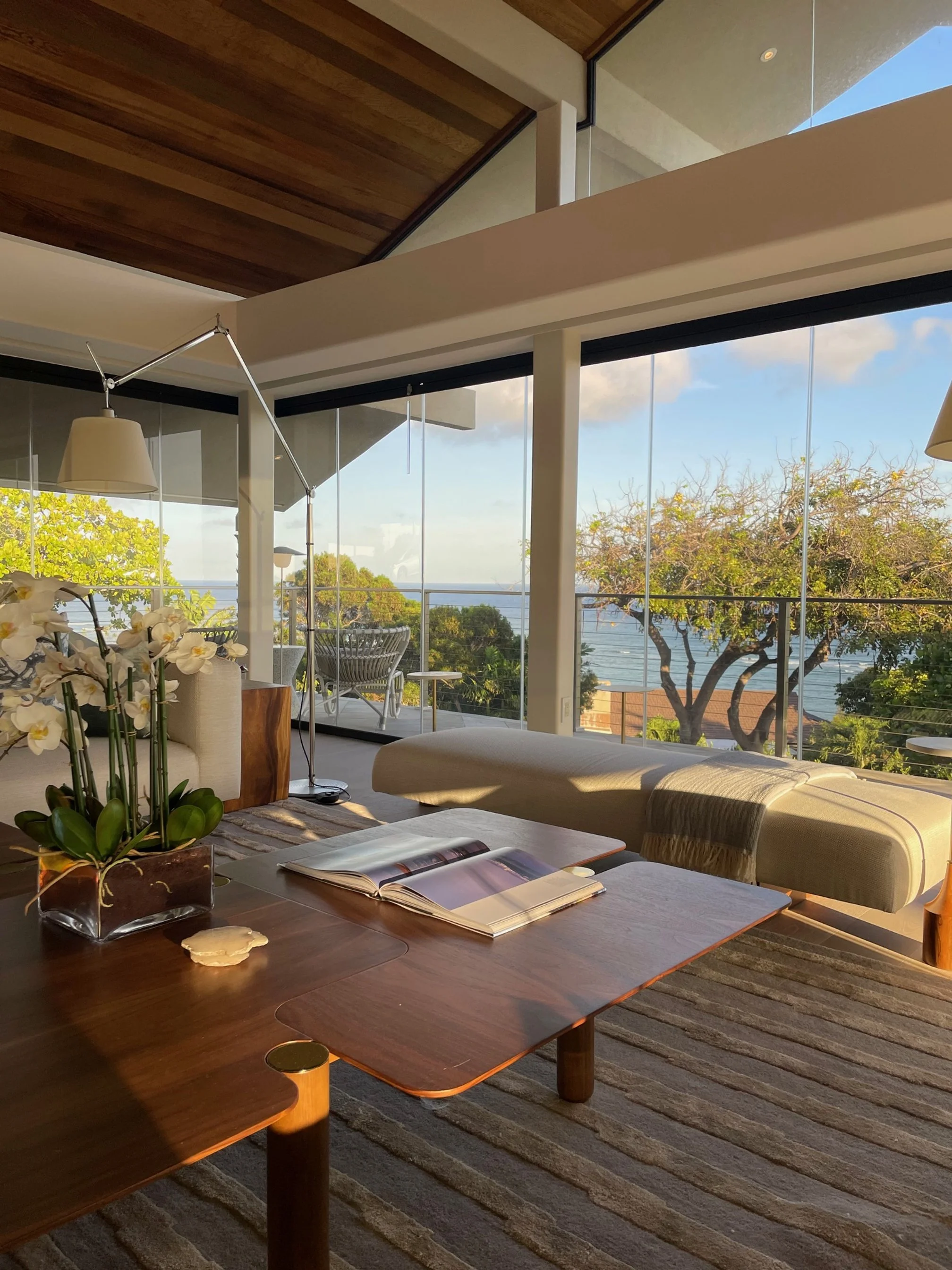 Modern living room with large glass windows overlooking trees and ocean, wooden ceiling, and a wooden dining table with an open magazine and a vase of white orchids.